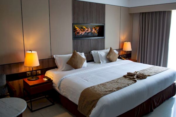 A cozy hotel room featuring a large bed, bedside lamps, brown accents, and a modern decor with a wall-mounted faux fireplace.