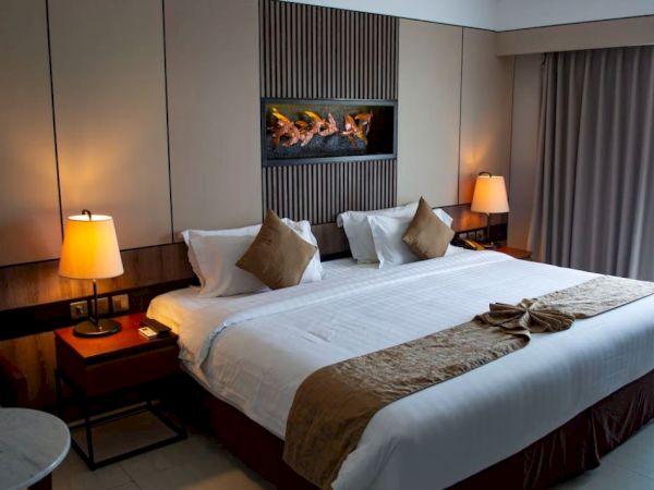 A cozy hotel room featuring a large bed, bedside lamps, brown accents, and a modern decor with a wall-mounted faux fireplace.