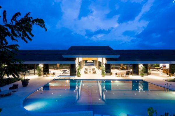 A luxurious building with a wide, illuminated swimming pool at dusk, surrounded by seating areas and lush greenery, under a blue sky.