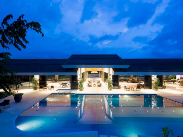 A luxurious building with a wide, illuminated swimming pool at dusk, surrounded by seating areas and lush greenery, under a blue sky.