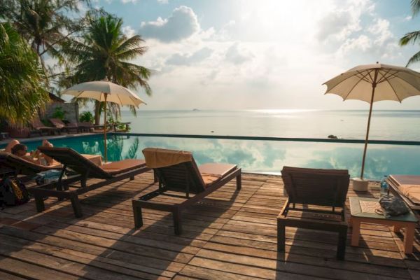 A serene poolside scene with lounge chairs, umbrellas, and a view of the ocean under a cloudy sky backed by sunlight.