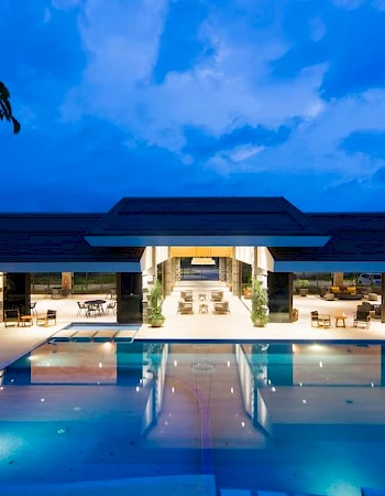 A luxurious open-air building with a large illuminated pool at night, surrounded by seating, under a deep blue sky.