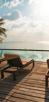 A serene poolside view with lounge chairs, umbrellas, and palm trees, overlooking a calm ocean under a bright sky.