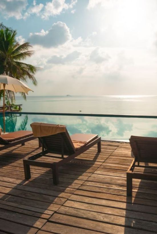 A serene poolside scene with lounge chairs, umbrellas, and a view of the ocean under a partly cloudy sky.