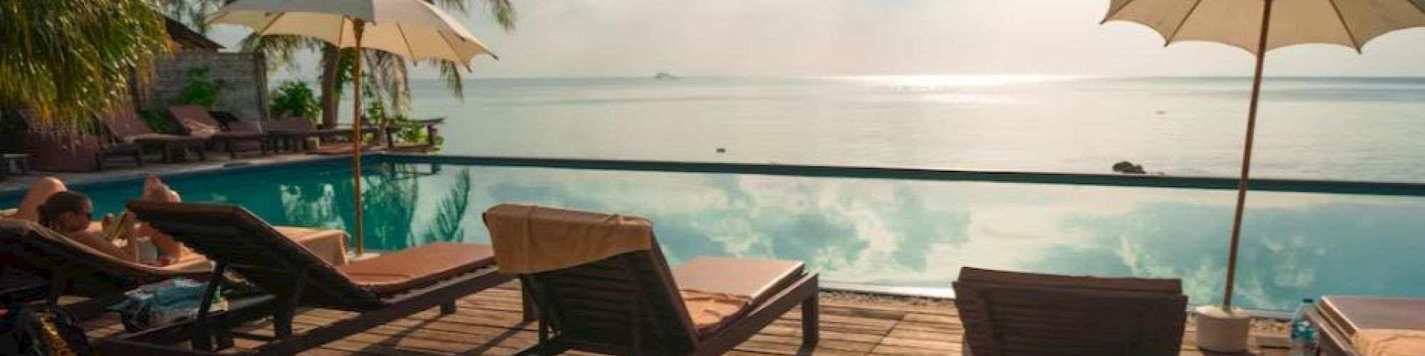 A serene poolside scene with lounge chairs and umbrellas overlooking a calm body of water, surrounded by palm trees under a sunny sky.