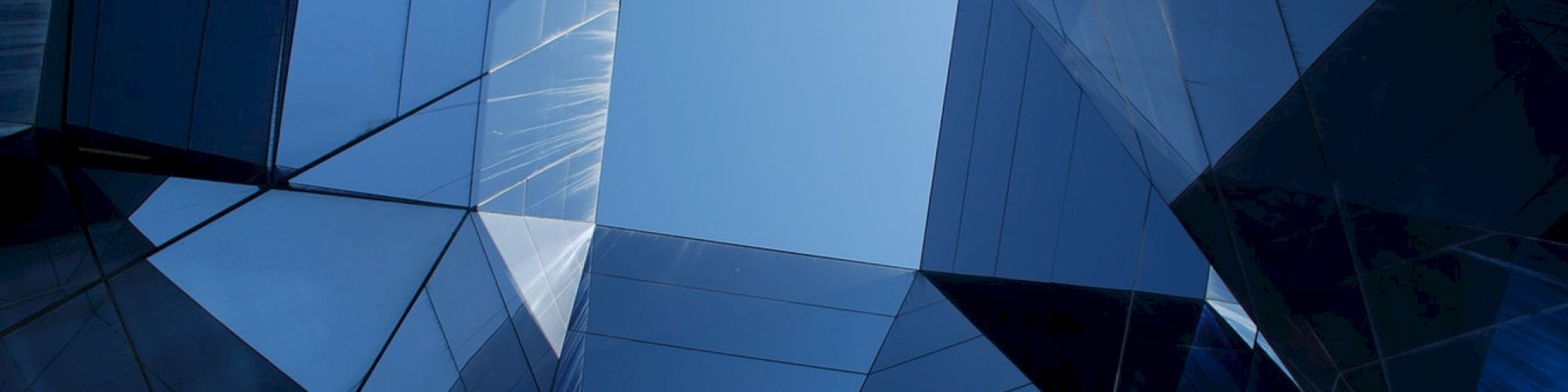 The image shows an upward view of modern glass buildings, forming geometric patterns against a clear blue sky.