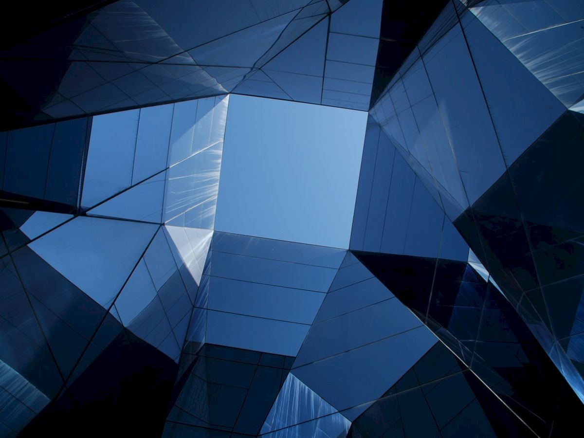The image shows a view upward through a geometric structure of reflective glass panels, creating a complex pattern against the blue sky.
