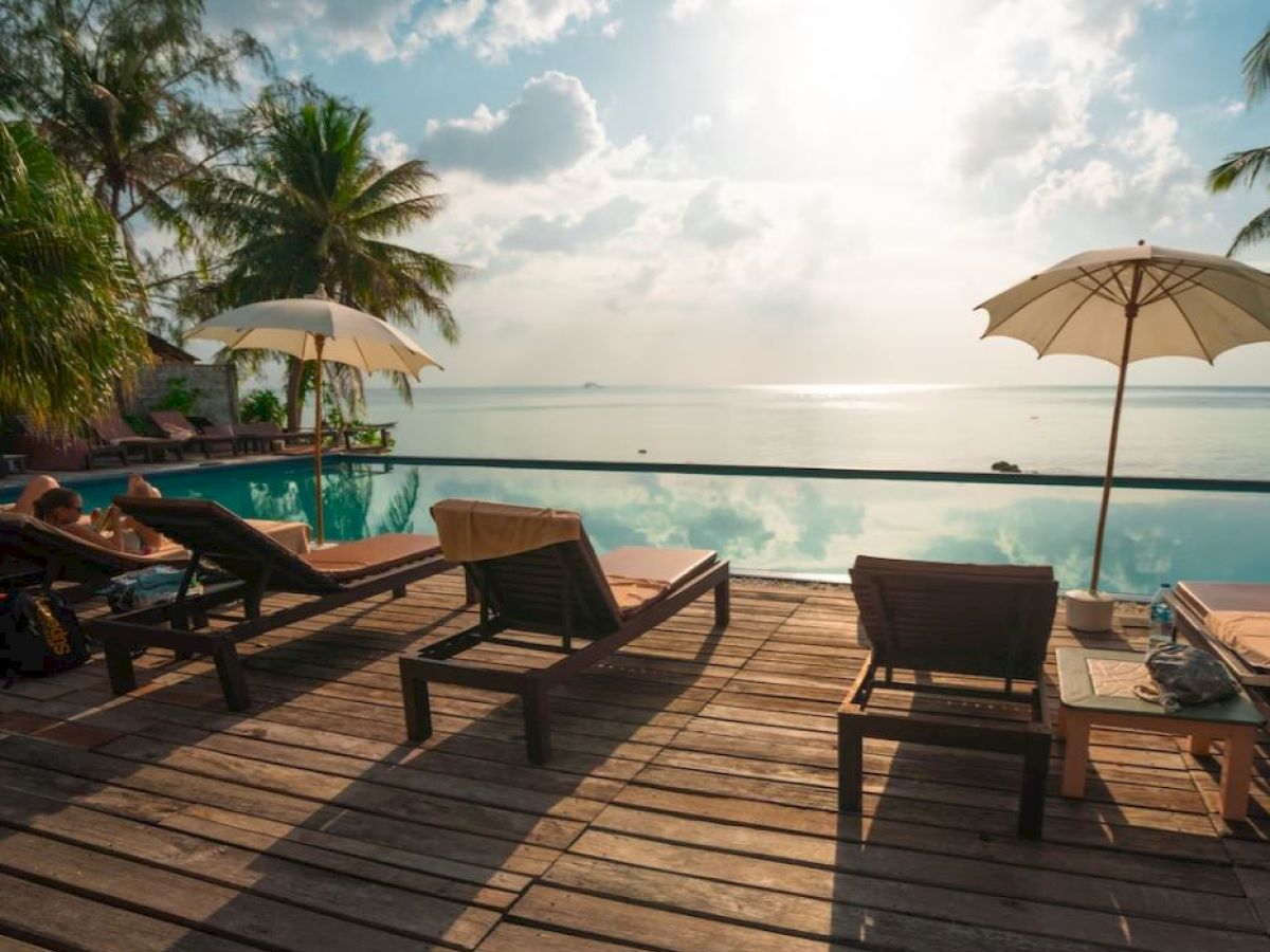 A serene poolside scene with lounge chairs, umbrellas, and palm trees overlooking calm ocean waters under a partly cloudy sky.