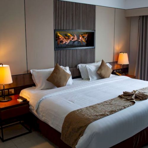 A hotel room features a large bed with pillows, warm lighting from bedside lamps, and a photo of a fireplace on the wall.