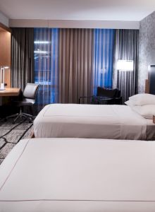 The image shows a modern hotel room with two beds, a desk, a TV, and stylish decor.