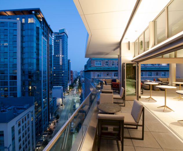 A modern rooftop terrace with seating overlooks a city street lined with tall buildings at dusk, under a clear blue sky.
