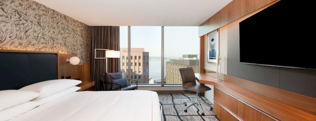 A modern hotel room with a large bed, TV, desk, chair, and window with an urban view is shown.