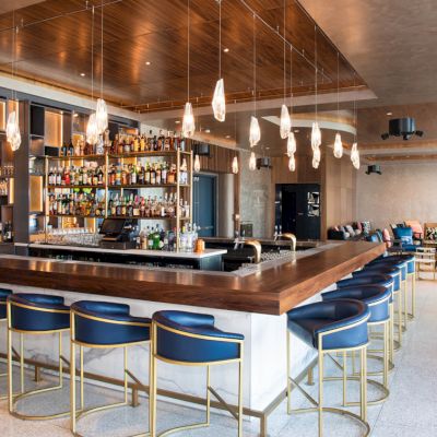 A modern bar with a sleek design features blue seating, a central liquor display, and hanging lights, creating a stylish atmosphere.