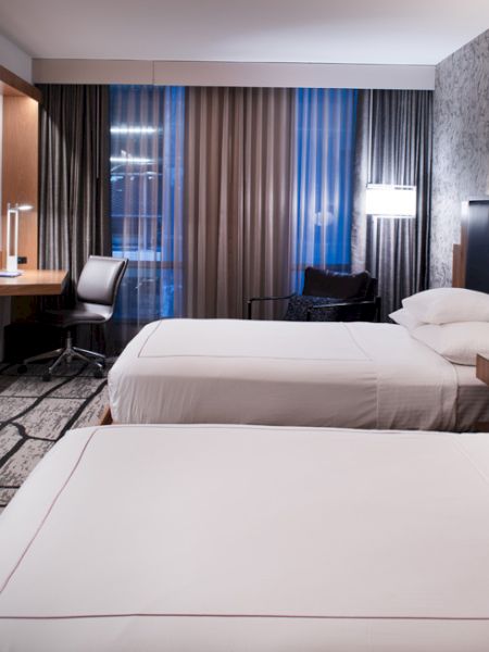 The image shows a modern hotel room with two beds, a TV, a desk, and a window with curtains. The decor is sleek and contemporary.