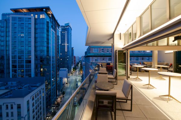 The image shows a modern cityscape with tall buildings and a sleek, illuminated balcony overlooking the street below at dusk.