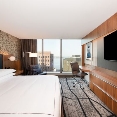 A modern hotel room features a large bed, wall-mounted TV, desk, chair, and a city view through floor-to-ceiling windows.
