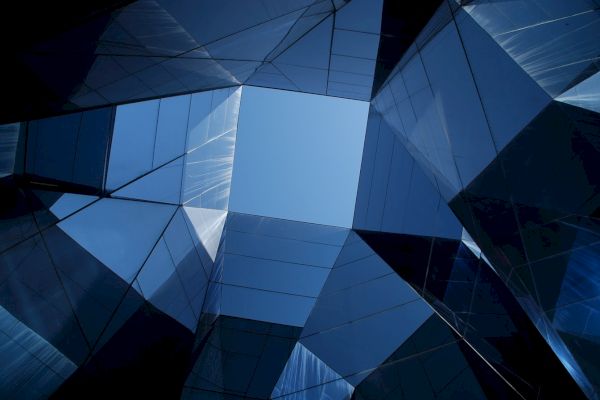 Looking up at a geometric arrangement of reflective glass and steel structures meeting at a clear blue sky.