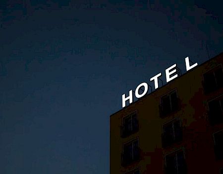 A building with a bright "HOTEL" sign on top, set against a dark, evening sky, with windows visible on the side wall.