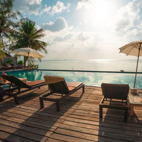 Lounge chairs and umbrellas by a pool overlook a calm sea, with palm trees and a bright sky in the background.