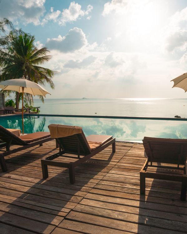 Lounge chairs and umbrellas by a pool overlook a calm sea, with palm trees and a bright sky in the background.