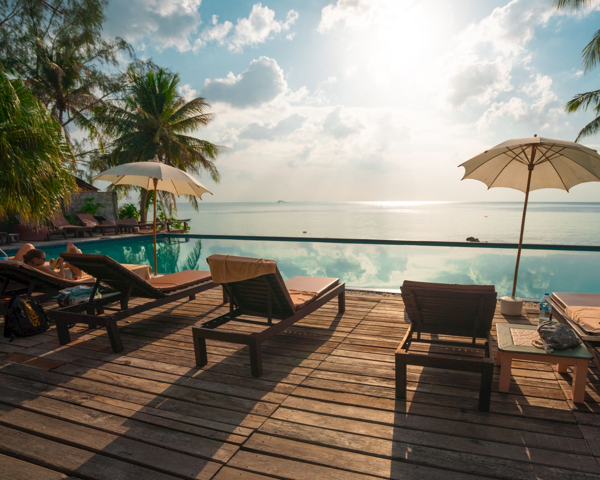Deck chairs and umbrellas overlook a pool with a serene ocean view and palm trees, creating a tropical paradise ambiance.