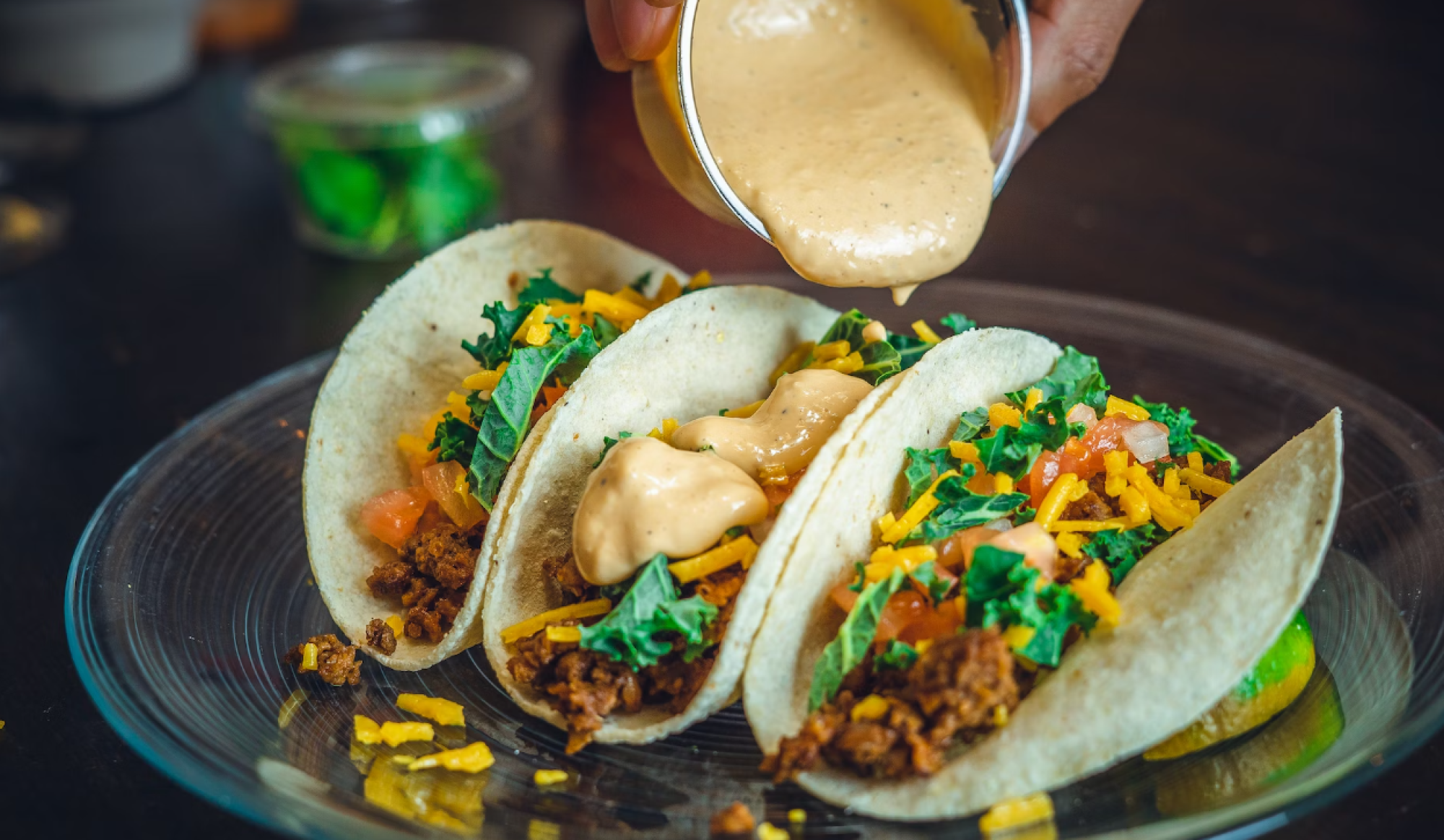 Three tacos filled with ground meat, cheddar cheese, lettuce, and tomatoes, topped with a creamy sauce being poured over them.