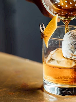 A cocktail being poured into a glass with ice and a lemon twist garnish, on a wooden surface.
