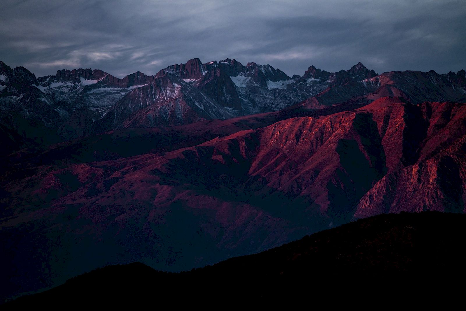 The image shows a mountain range with dramatic lighting, highlighting rugged peaks and ridges under a moody sky at sunset or sunrise.