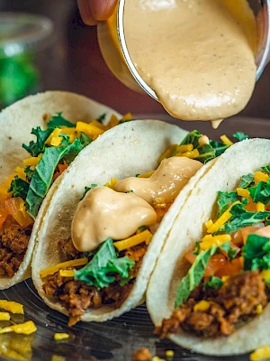 Three tacos filled with ground meat, lettuce, tomatoes, and cheese are being topped with a creamy sauce on a plate.