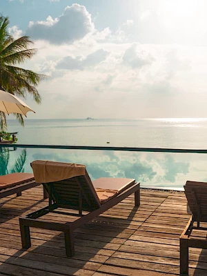 Lounge chairs and umbrellas by a pool overlook a tranquil sea, framed by palm trees under a partly cloudy sky, creating a serene setting.