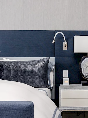 A modern bedroom with a navy blue headboard, white bedding, a nightstand with a lamp, and wall-mounted reading light.