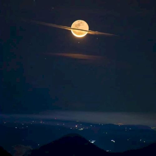 A glowing moon appears to have Saturn-like rings, with clouds creating an illusion. City lights and mountains are visible below in the night sky.
