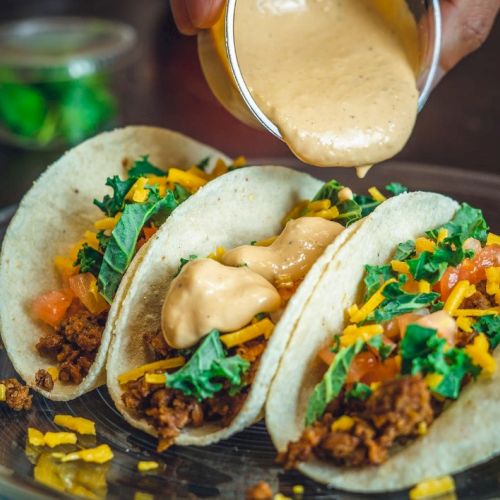 Three tacos filled with ground meat, shredded cheese, and greens are being topped with a creamy sauce.