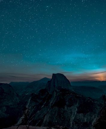 A starry night sky over a mountainous landscape with a glowing horizon and silhouetted trees in the foreground.