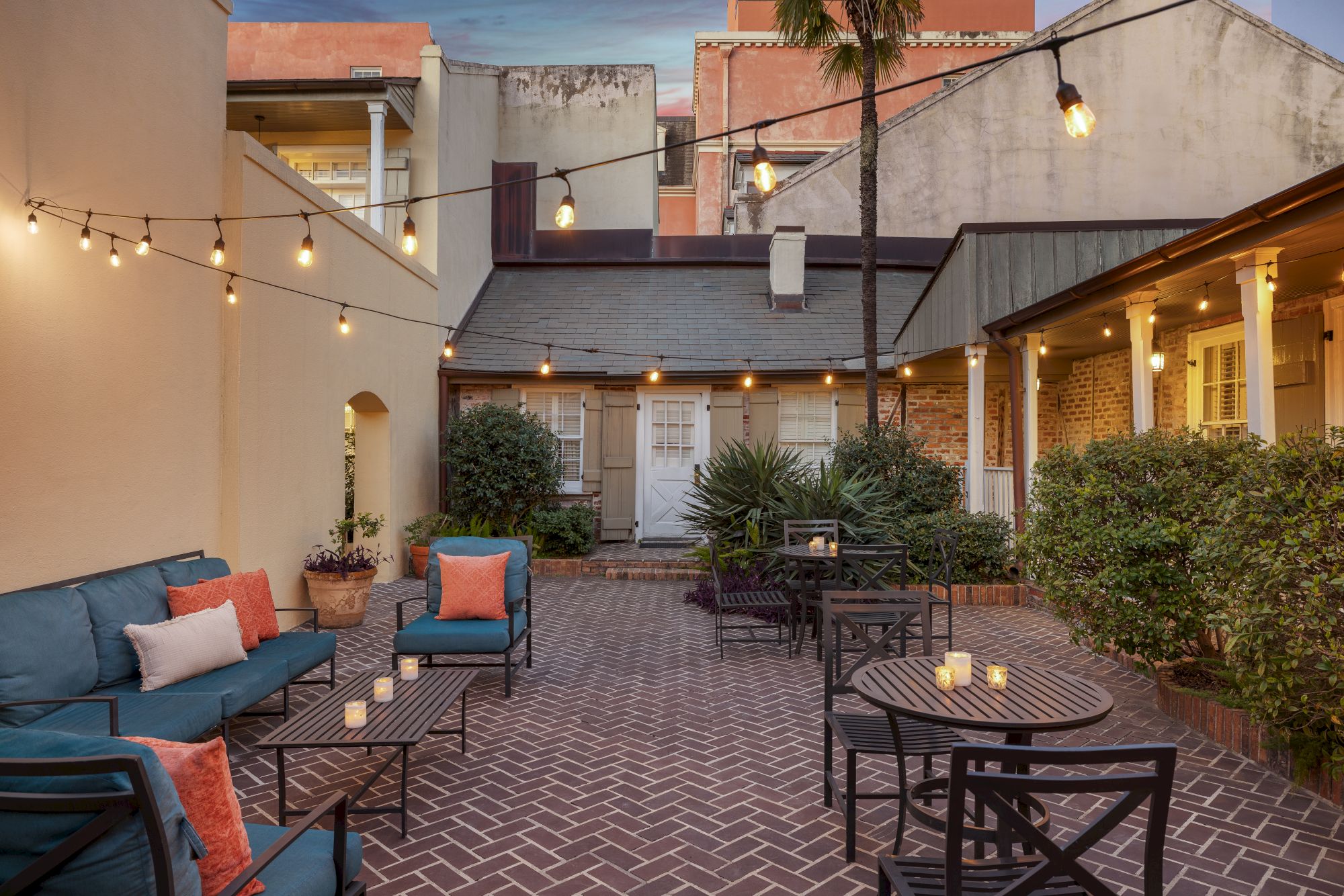A cozy courtyard with string lights, brick pavers, outdoor seating, cushioned chairs, and potted plants under a warm evening sky.