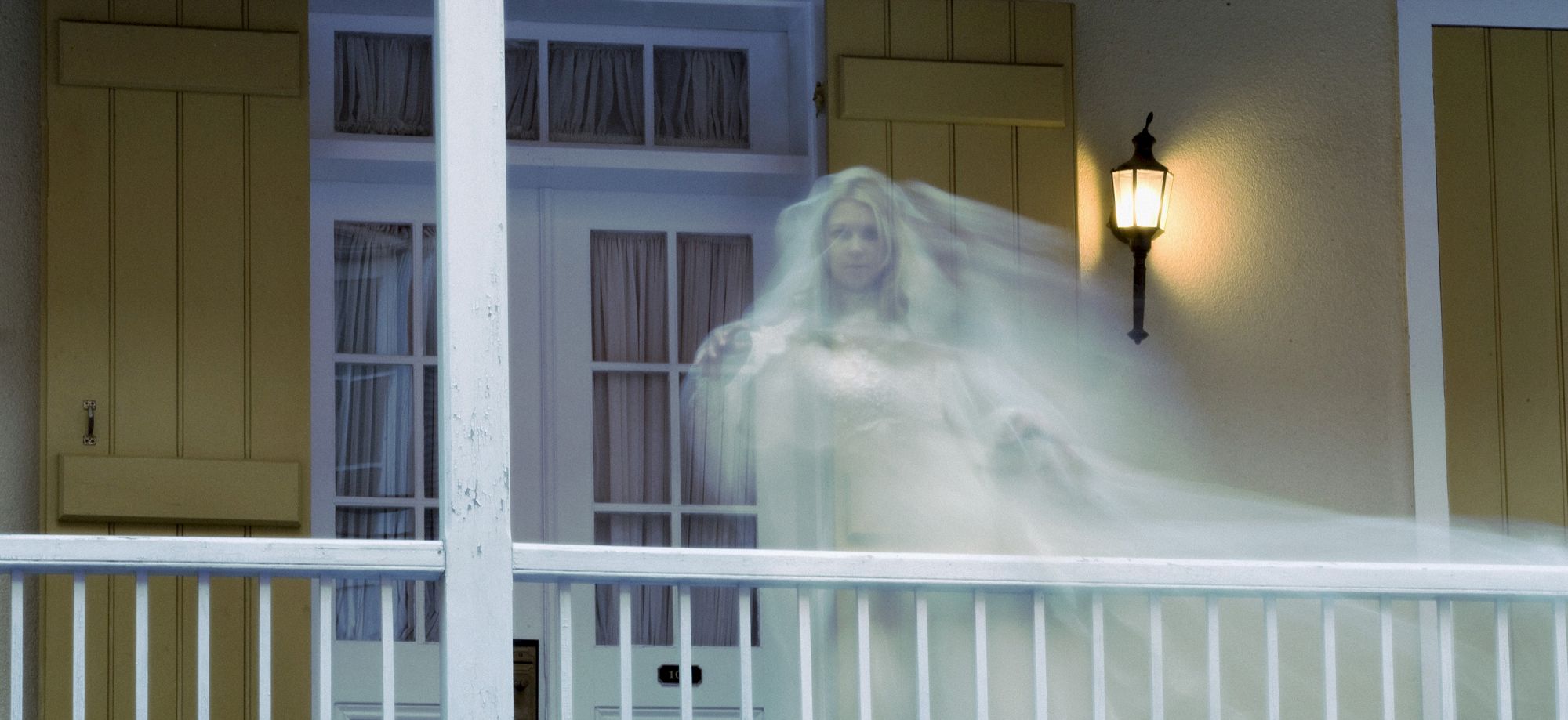 The image depicts a blurred figure in a white dress, resembling a ghostly apparition, standing on a porch near a lit lantern at night.