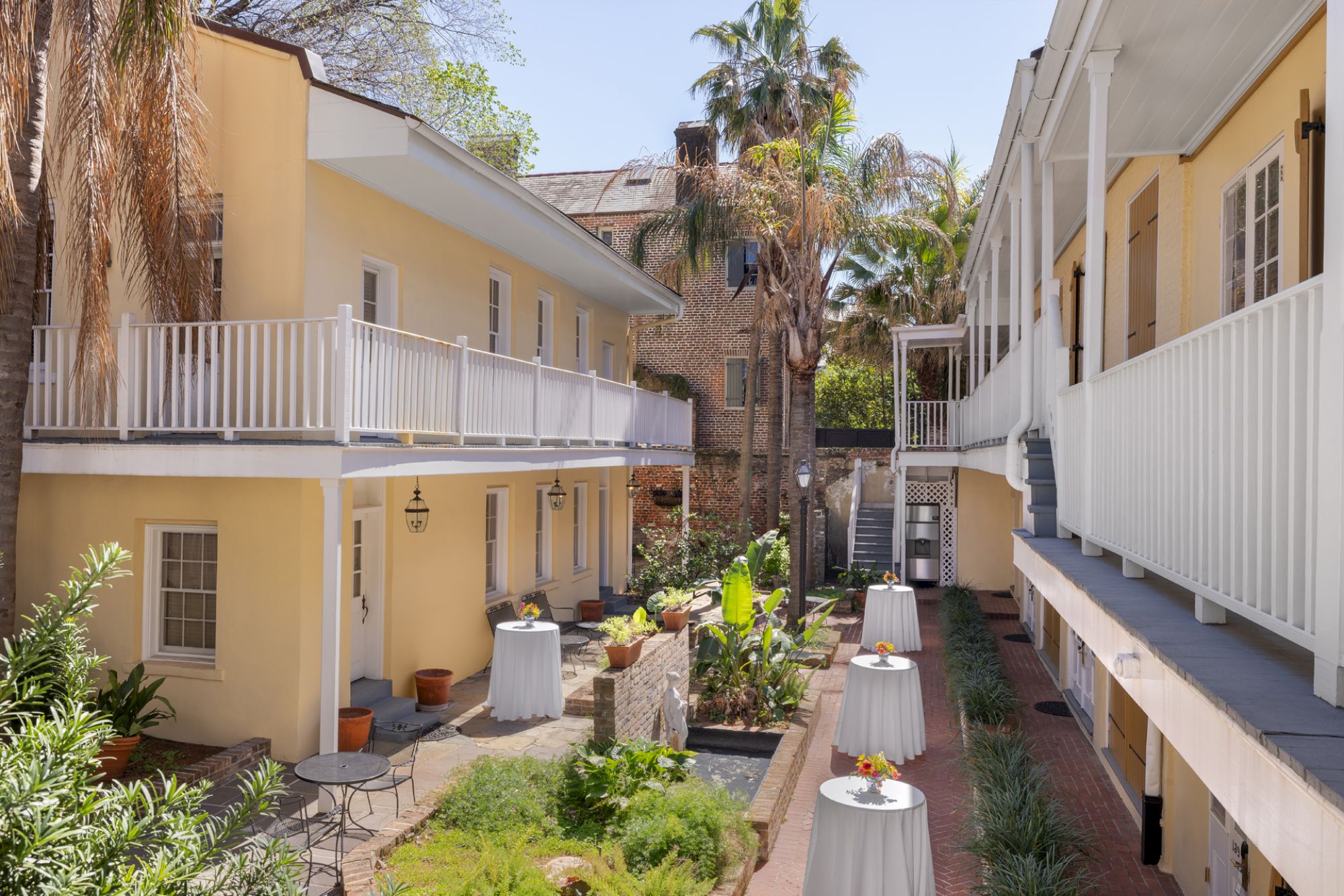 A courtyard with yellow buildings, a garden, and tables set with white cloths and flowers. Palm trees add a tropical touch to the scene.