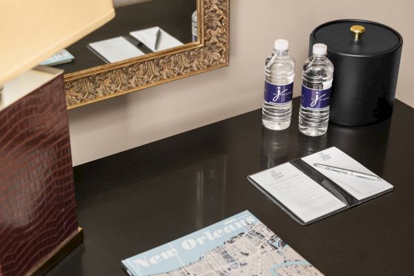 A desk with two water bottles, a black container, a hotel information folder with a pen, a magazine about New Orleans, and a decorative mirror.