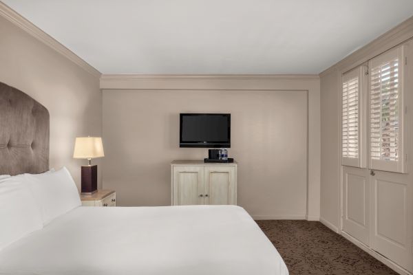 A hotel room with a bed, nightstand, lamp, wall-mounted TV, and a cabinet with bottled water on it, near windows with shutters.