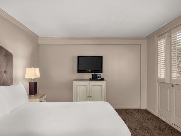 A hotel room with a bed, nightstand, lamp, wall-mounted TV, and a cabinet with bottled water on it, near windows with shutters.