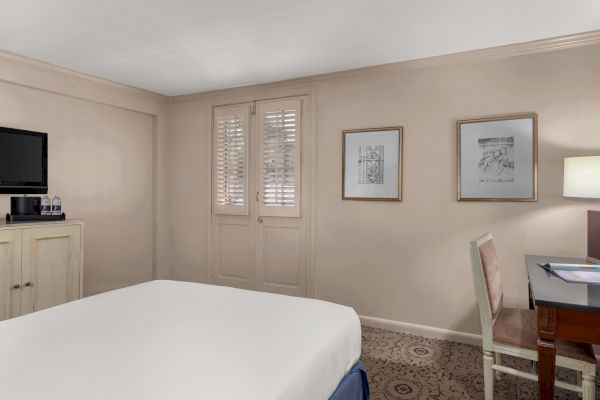 A hotel room with a bed, TV, desk, chair, paintings, door with blinds, lamp, and bottled water on a small cabinet.
