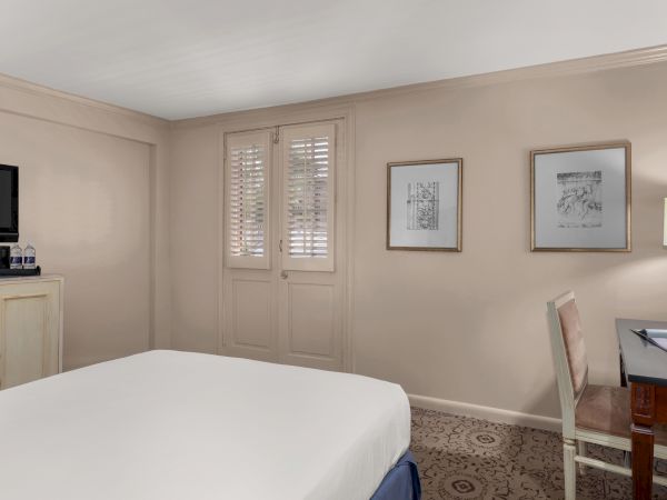 A hotel room with a bed, TV, desk, chair, paintings, door with blinds, lamp, and bottled water on a small cabinet.