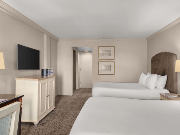 A hotel room with two beds, a TV, lamps, a dresser with water bottles, and wall art. The room is clean and simply decorated.