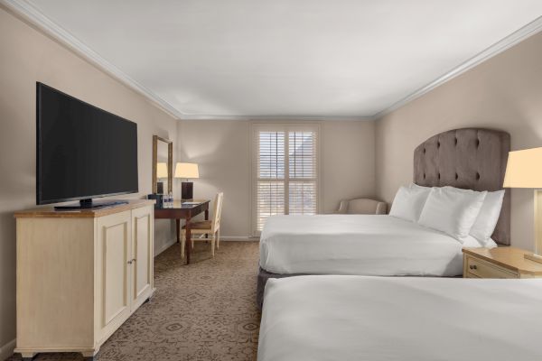 The image shows a hotel room with two beds, a large TV, a desk, a chair, a lamp, and a window with shutters.