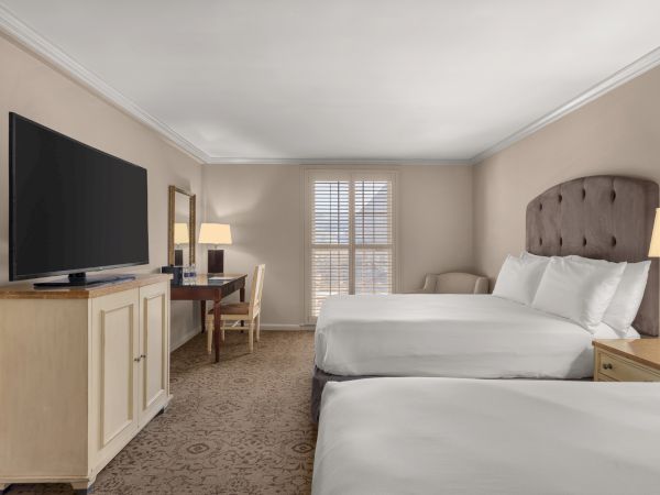 The image shows a hotel room with two beds, a large TV, a desk, a chair, a lamp, and a window with shutters.
