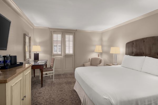 A hotel room with a large bed, TV, desk with chair, armchair, lamps, and bottled water atop a cabinet.