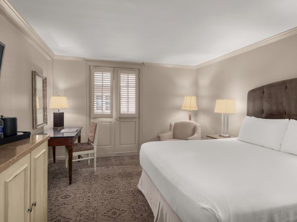 A hotel room with a large bed, TV, desk with chair, armchair, lamps, and bottled water atop a cabinet.