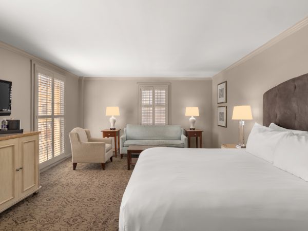 A hotel room with a bed, armchair, sofa, TV, lamps, and a window covered by blinds.