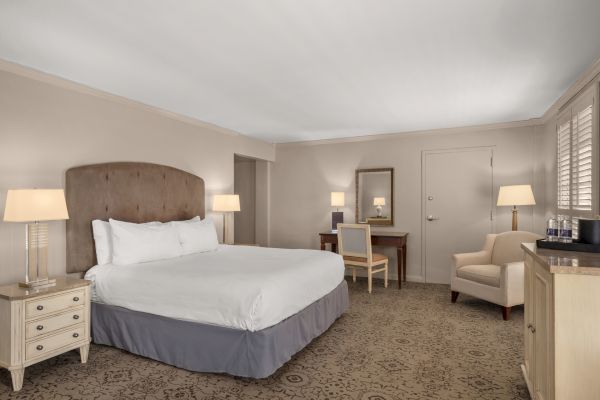 A hotel room with a bed, nightstands, lamps, a desk with a mirror, a chair, an armchair, and a cabinet.