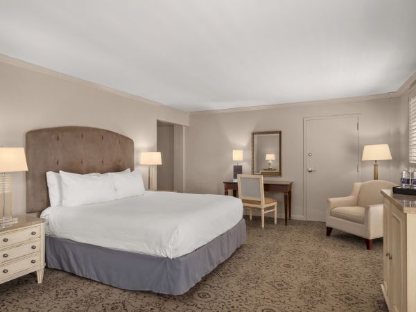 A hotel room with a bed, nightstands, lamps, a desk with a mirror, a chair, an armchair, and a cabinet.